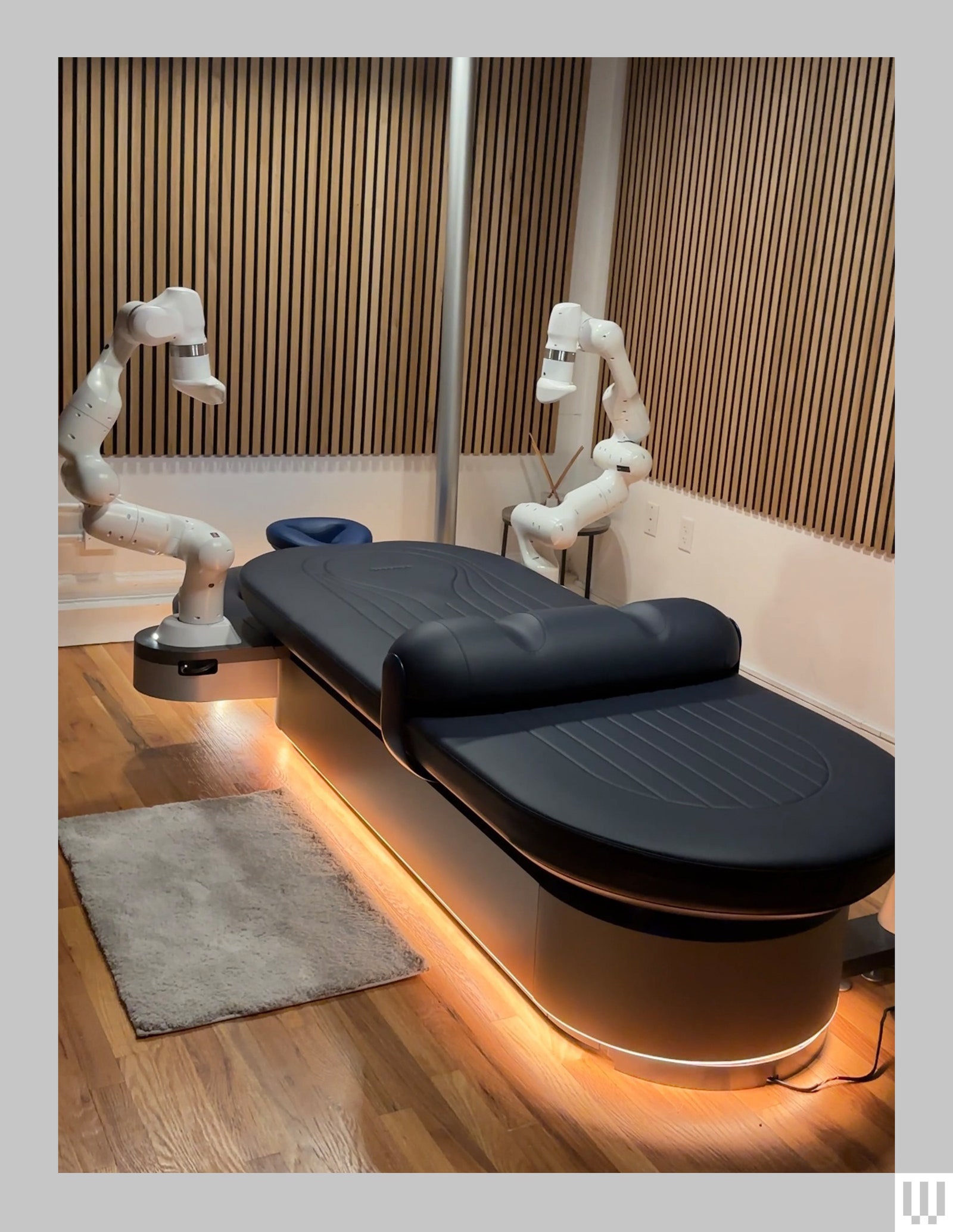 Black padded massage table in the center of the room with two angular white robotic arms hovering over the left and...